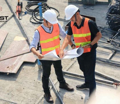 Two construction workers in safety vests and helmets reviewing blueprints on a building site, with metal rods and construction materials in the background