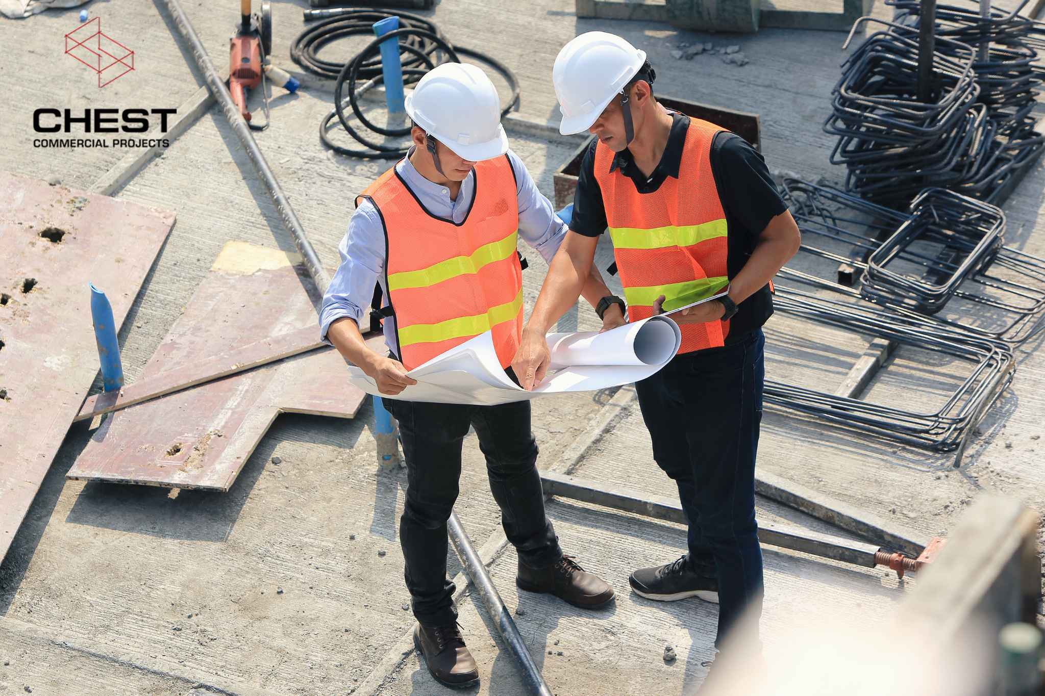 Two construction workers in safety vests and helmets reviewing blueprints on a building site, with metal rods and construction materials in the background