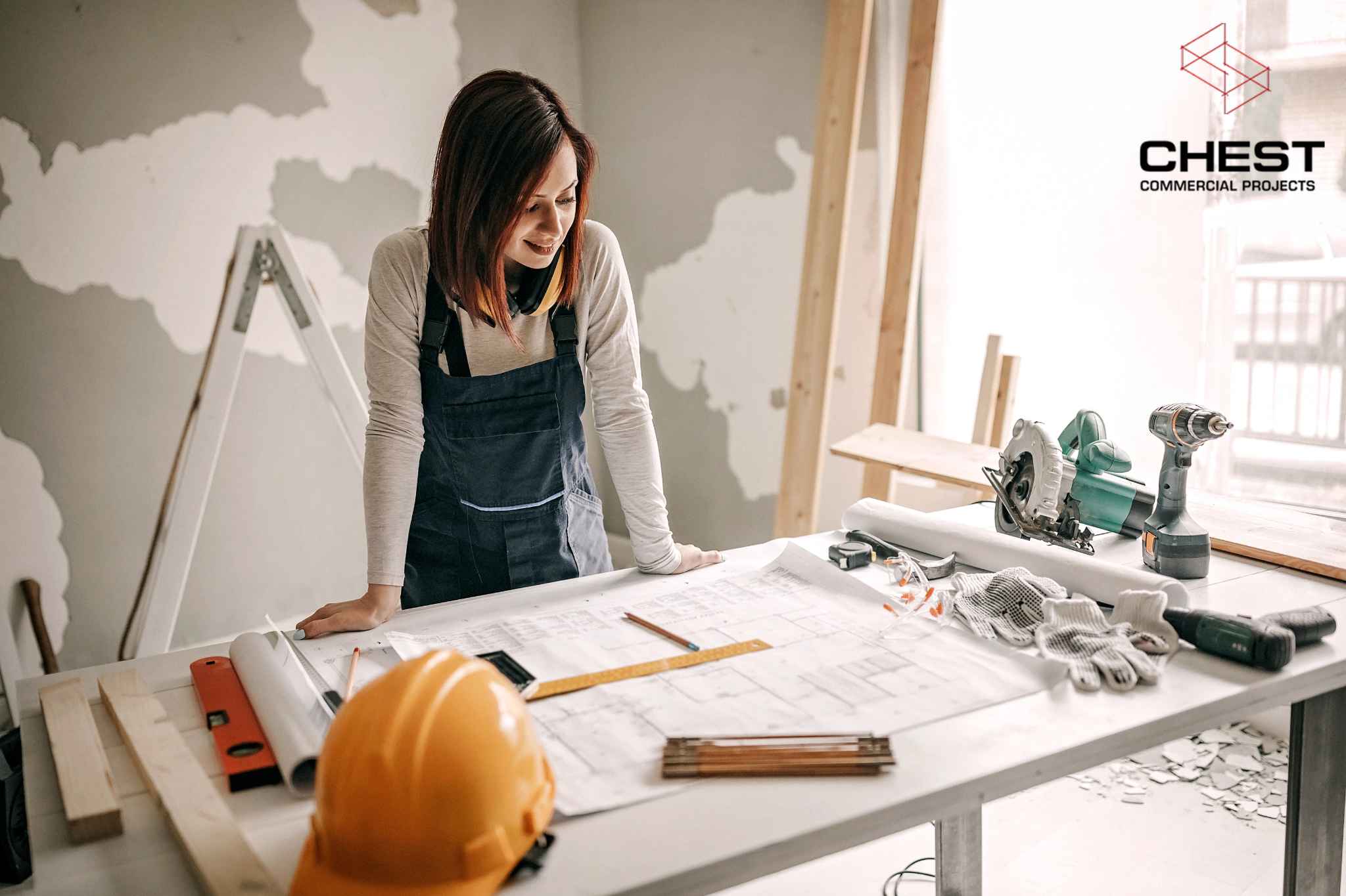 Female contractor reviewing office fitout floor plans during a commercial fitout or renovation project in Perth, with tools, safety helmet and Chest Commercial Projects branding.