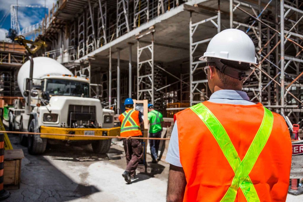 Construction worker in high-visibility safety gear overseeing a busy commercial construction site with scaffolding, workers and heavy equipment.